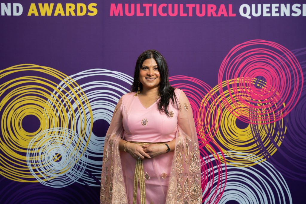 Sara Shams dressed in a beautiful traditional pink sari standing in front of the QLD Multicultural awards banner